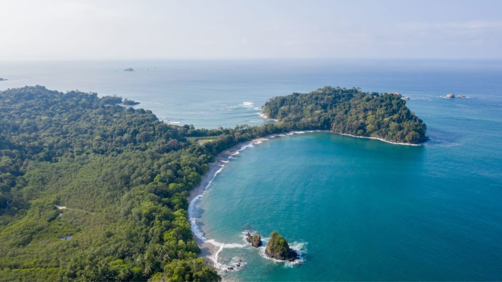 Aerial view of a lush green coastal landscape in Costa Rica, with a curving bay and small rocky islets, exemplifying the stunning beauty of some of the best beaches in Costa Rica beneath a clear sky.
